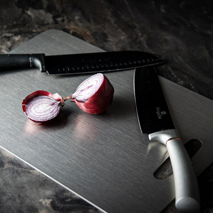 Knightsax Privateer 
®
 medical-grade titanium cutting board, silver, with two knives and a red onion.